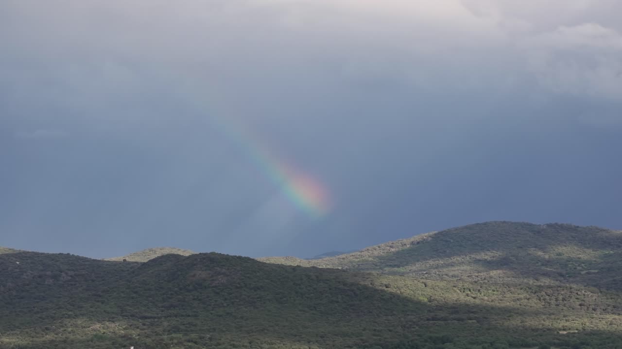 Cinematic 70mm drone shot across forested mountains where the treetops sparkle in the sun, revealing one arm of a rainbow arc in the sky and creating a magical, atmospheric sunburst view