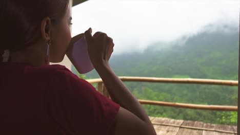 asian girl sits on balcony drinks tea from cup and enjoyes beautiful view of the mountains