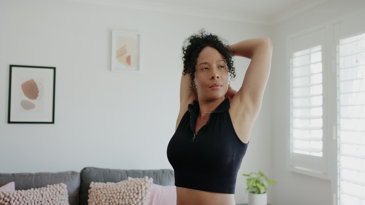 Eine afroamerikanische Frau in Crop-Top und Leggings streckt die Arme, bindet einen Dutt und wärmt sich am Sofa auf.