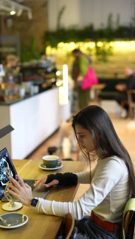 una mujer trabajando en una computadora portátil en un café, posiblemente revisando imágenes médicas.