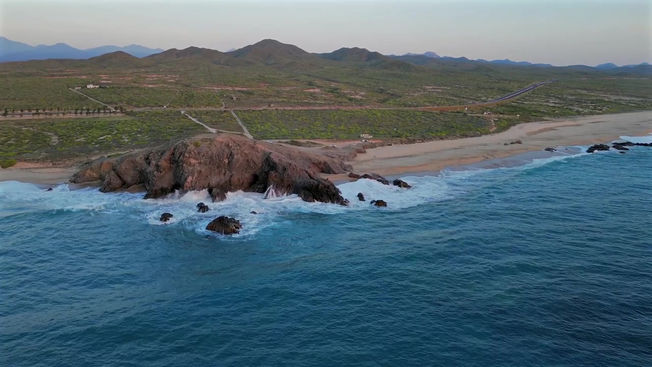 Drone avança sobre águas calmas do oceano em direção à ponta rochosa da Praia Secret Rocks, abaixo de Todos Santos, com ondas quebrando contra a ponta e um carro passando por uma estrada distante, enquanto o deserto verde