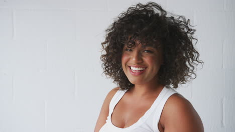retrato de una mujer sonriente y vestida casualmente en un chaleco de pie contra la pared del estudio de ladrillo blanco