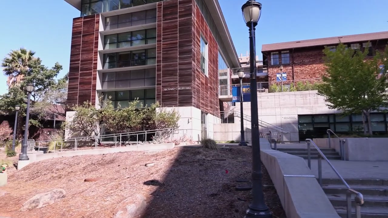 Perspectiva pessoal caminhando para trás, revelando a arquitetura moderna do Richard C. Blum Hall e os edifícios circundantes no campus da Universidade da Califórnia, Berkeley.