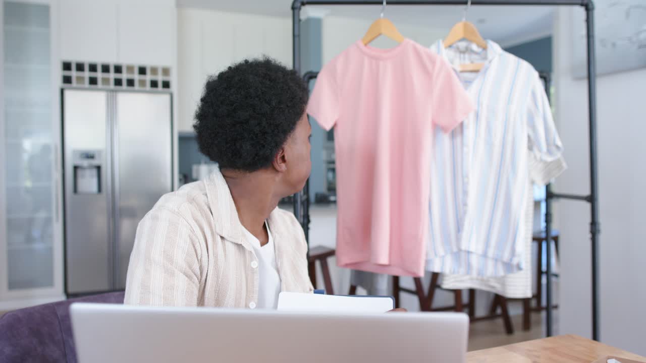 Un homme assis travaillant à distance sur son ordinateur portable, jetant un coup d'œil à un portant et soulevant l'ourlet d'un t-shirt rose pour vérifier la taille.