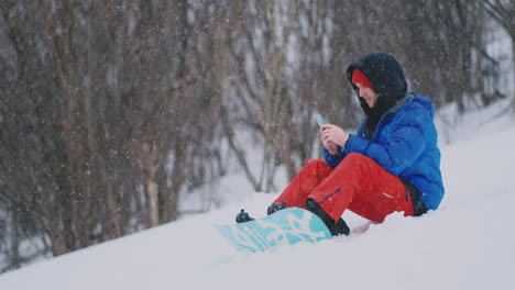 un snowboarder sentado en la nieve toma fotos por teléfono de un hermoso paisaje turístico para las redes sociales. blogger en el resort. escribir mensajes de texto a amigos usando su teléfono inteligente