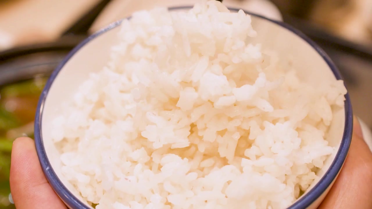 Close-up of hands using black chopsticks to eat steamed jasmine rice from a bowl.