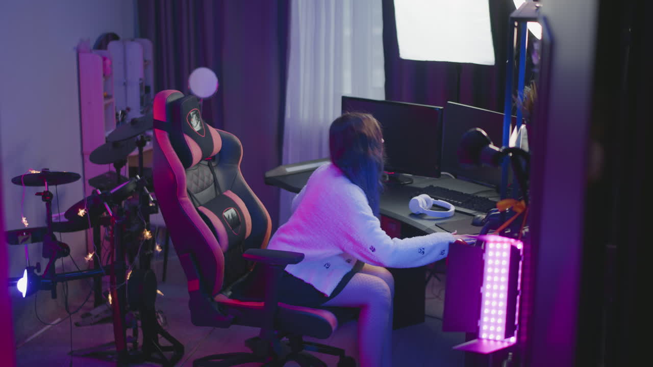 Young girl in fluffy sweater stands behind gaming chair in neon lit studio adjusts seat at desk with computer monitors ring light and white headphone on table then sits under ambient pink purple glow