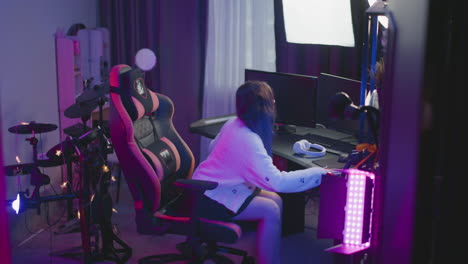 young girl in fluffy sweater stands behind gaming chair in neon lit studio adjusts seat at desk with computer monitors ring light and white headphone on table then sits under ambient pink purple glow
