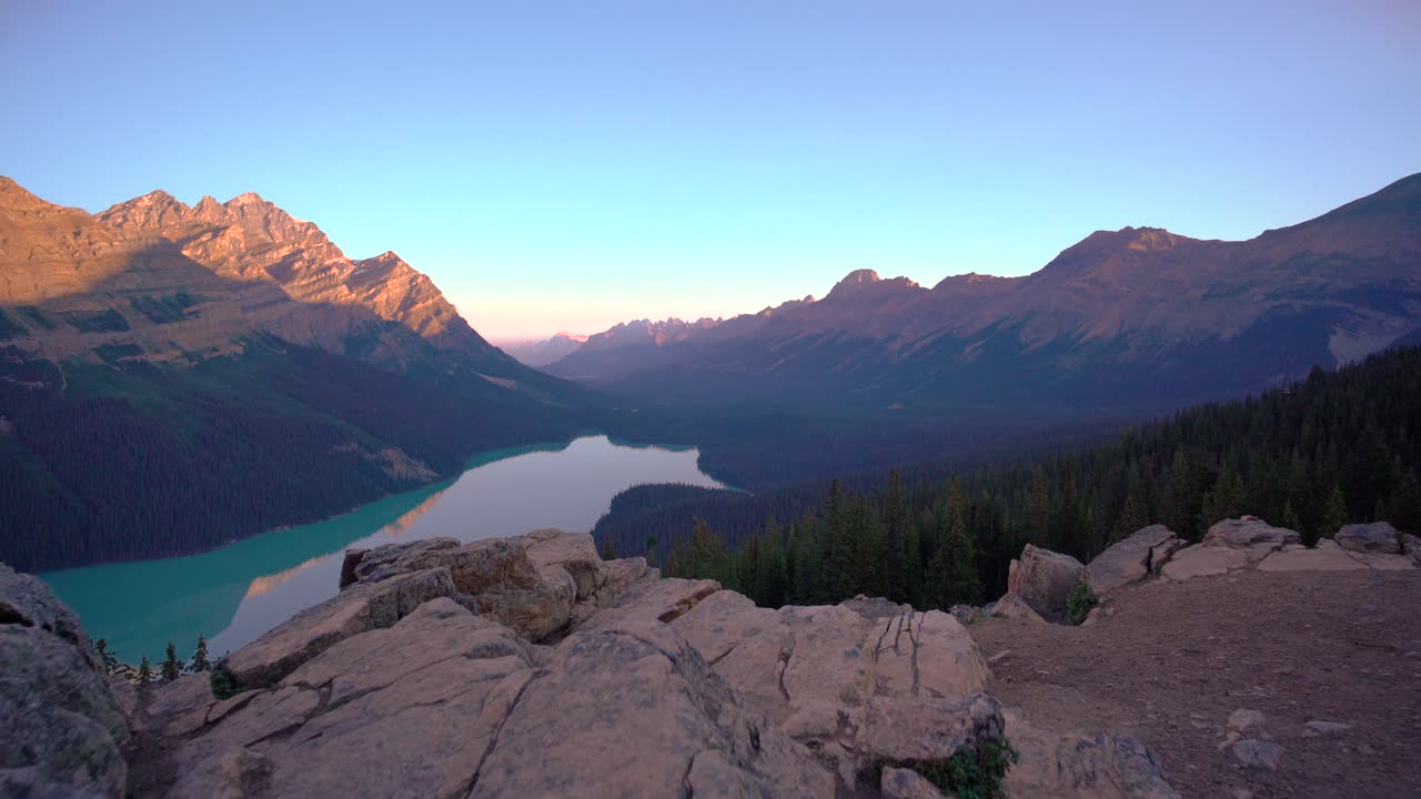 Drone glides over turquoise lake nestled between pine-covered slopes and majestic mountain ranges in Banff National Park. Morning light casts warm tones across rugged terrain and peaceful water.