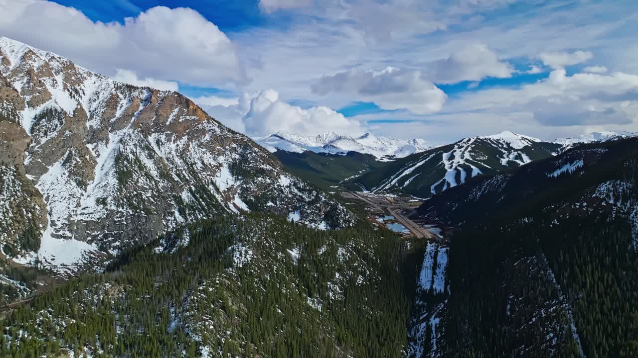 Flying over rocky valley in Frisco Gulch with thick pine trees and morning sun, aerial pullback with highway in distance, snow spring on mountains