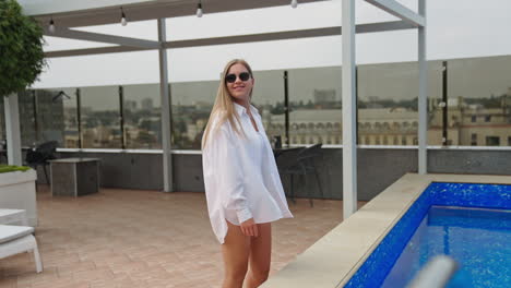 young blonde lady in white swimsuit and shirt walks near the swimming-pool. relaxing lounge zone at the roof of a building. cityscape at backdrop.