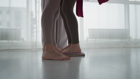 close up of two women standing side by side in yoga studio as one rests her feet firmly on nail mat while other stands barefoot beside her, emphasizing contrast between grounding practices