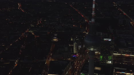 Luces-Parpadeantes-De-La-Ciudad-Por-La-Noche-Con-Rascacielos-De-La-Torre-De-Televisión-En-Primer-Plano,-Paisaje-Urbano-De-Drones-Aéreos