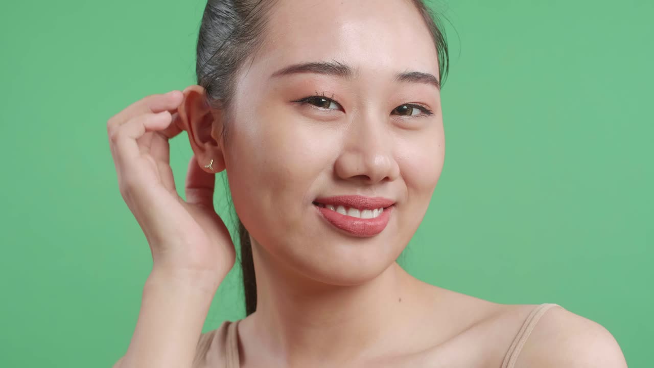 스튜디오에서 녹색 화면 배경에서 카메라에 미소 짓는 동안 머리카락을 고치고 얼굴을 만지는 아시아 여성의 클로즈업