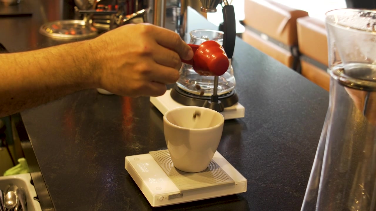 Primo piano di chicchi di caffè interi aggiunti a una tazza bianca su bilancia digitale al bancone di una caffetteria specializzata