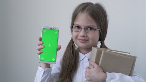 schoolgirl in white school blouse and glasses holding books and shows phone with green screen mock up. chroma key green screen smartphone set up for advertising. education, technology, gadget and kid