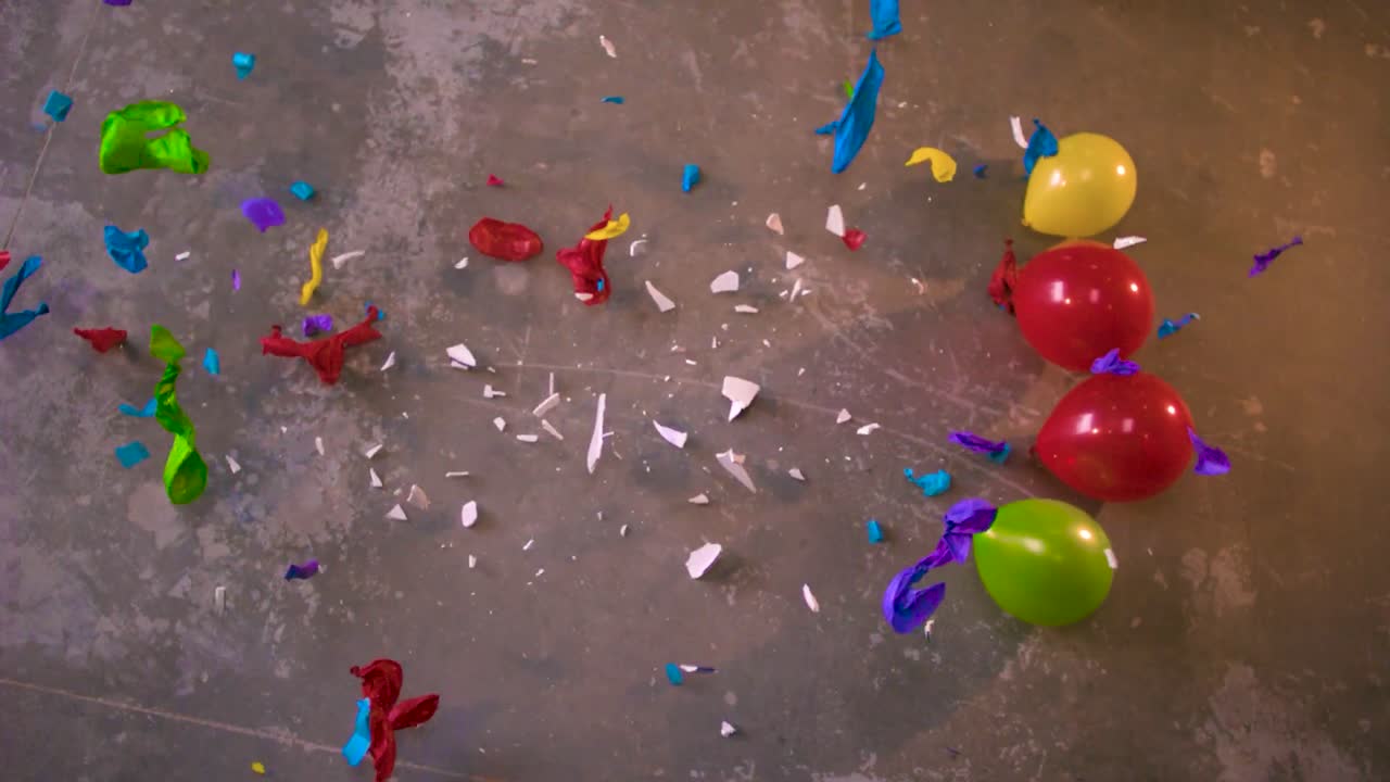 A plate being dropped in the middle of colorful balloons and it breaks in pieces and the bits and shards shatter and make balloons explode around it in slow motion. view from the top, concrete floor.