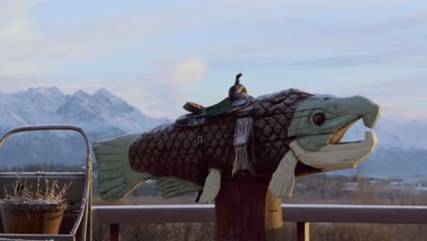 Amanecer-En-Alaska,-Panorámica-Alrededor-De-La-Estatua-De-Trucha-De-Madera-Con-Silla-De-Montar-Que-Revela-Hermosas-Montañas-A-Distancia-De-Anclaje-4k