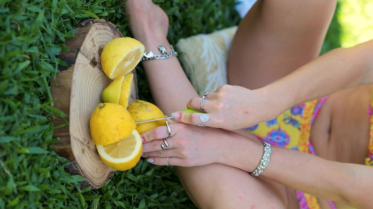 Modelo femenina de joyería cortando limones en un día soleado y cálido al aire libre en el césped cerca de la piscina. Video vertical.