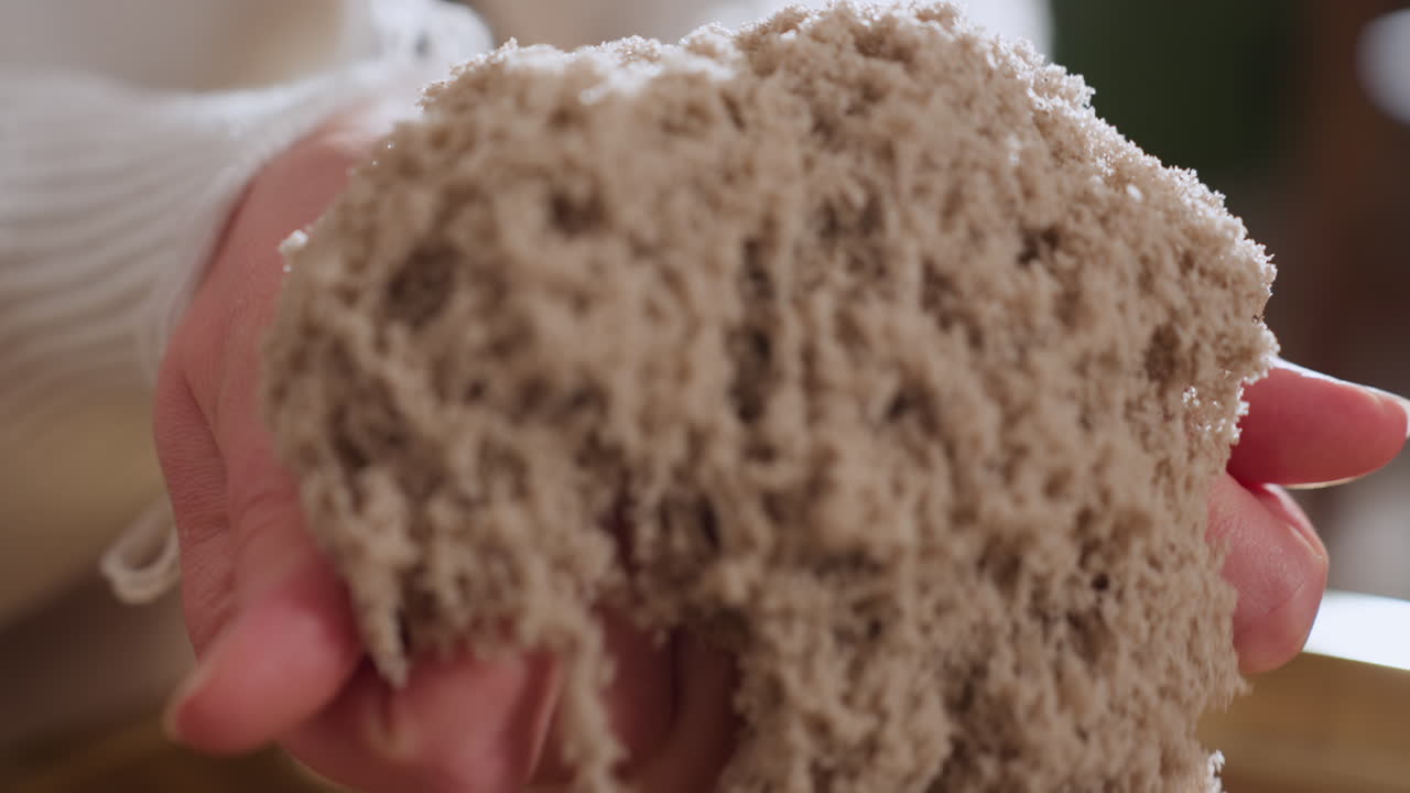Close up of visitor gently lifting soft wet soil mold with both hands during calming sensory therapy session, capturing tactile experience of textured material on golden tray