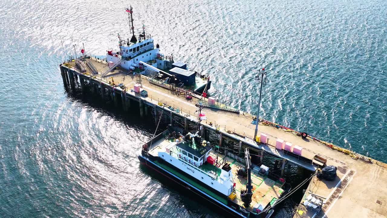 Drone captures fishing vessel at pier in bright daylight, smooth lateral movement, coastal industrial setting
