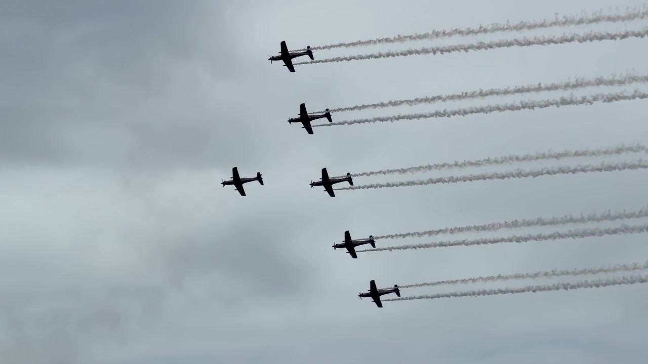 Silhouetted vintage warbirds perform formation maneuvers with white smoke trails against an overcast sky.