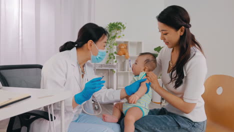 children vaccination concept, smiling female doctor or nurse standing in medical holding syringe in hands with mother and little boy in background. pediatrician vaccinating little baby in clinic