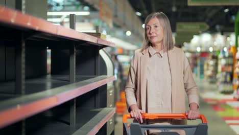 senior woman shopper in supermarket, grocery store near empty shelves counters. elder mature female panic buying and stockpiling. groceries supplies with sold out products.