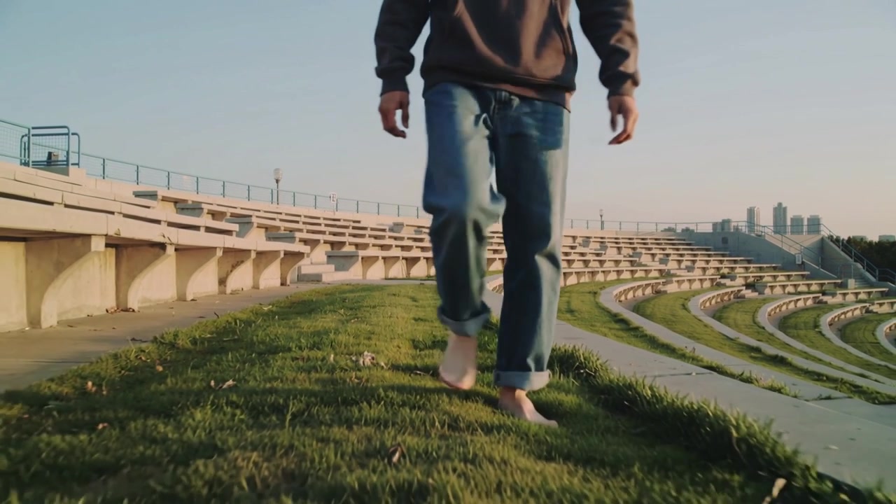 Uomo che entra nell'inquadratura, fa un passo a piedi nudi e cammina in equilibrio su un crinale erboso al parco, indossando jeans