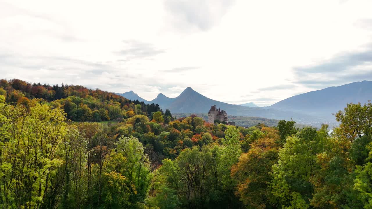 Autumn forests surround Burg Menthon-Saint-Bernard, nestled among rolling hills with a distant mountain range and lake creating landscape blending natural beauty and historic architecture in France
