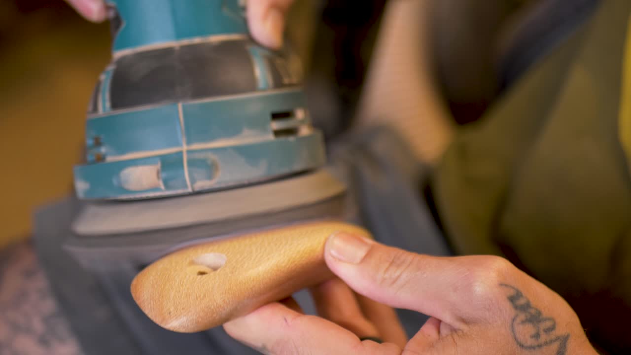 Slow motion close-up of man with hand tattoos using electric orbital grinder sanding power tool to shape wooden block for indoor climbing holds at shed garage warehouse in Australia industry carpentry