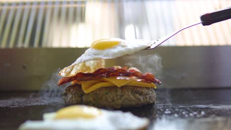 placing egg on top of a burger with cheese and bacon on the grill smoke slow motion