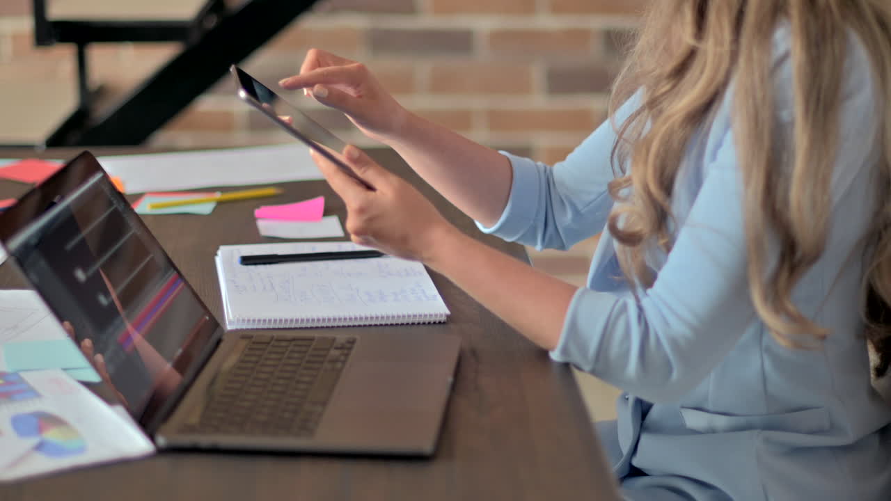 Woman in blue blazer working on a tablet in front of a laptop, at a desk, in an office