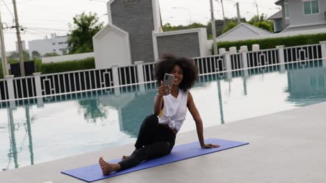 a female dressed for exercise sits on a yoga mat talking smart phone video call.