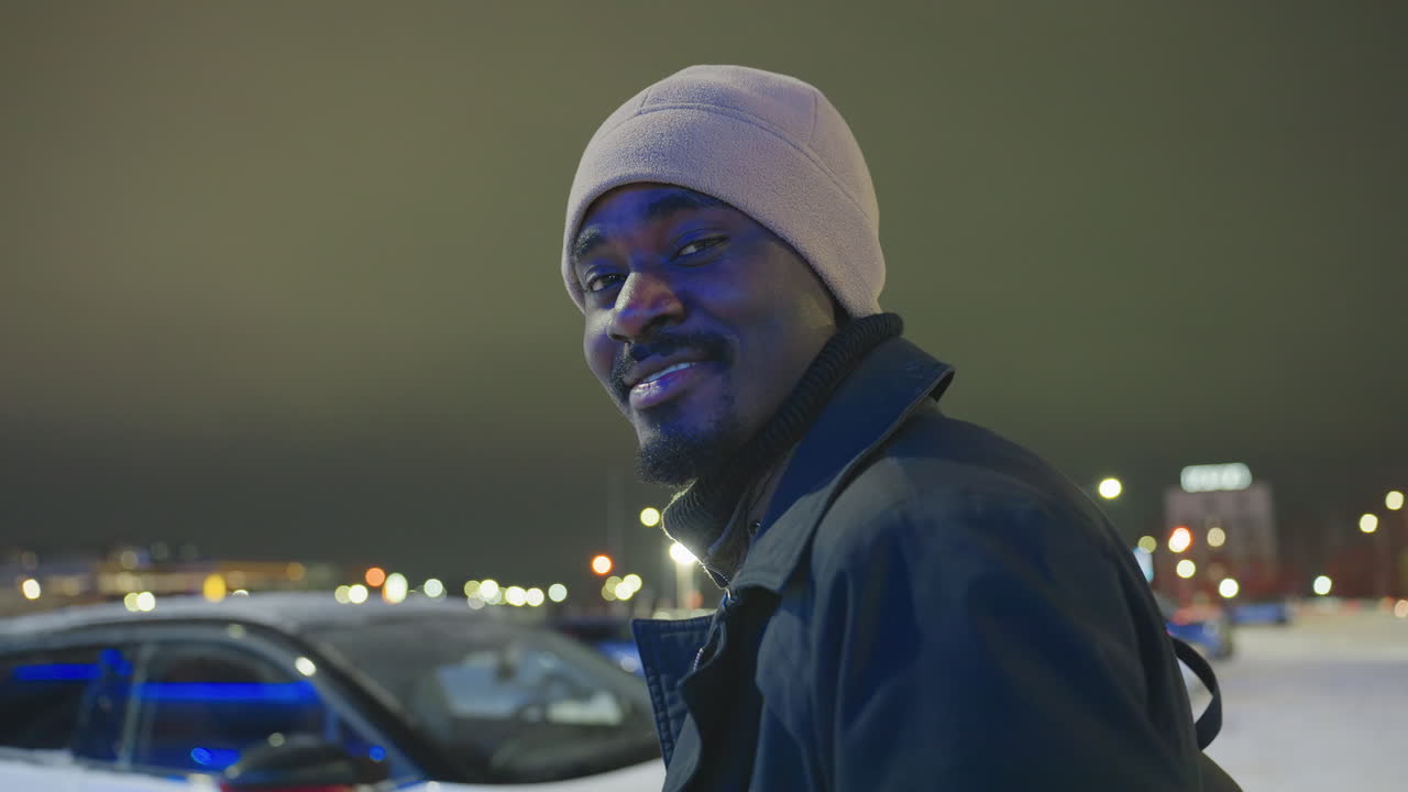 Man wearing warm coat and hat turns with gentle smile while standing outside on cold winter night, illuminated by blurred city lights creating relaxed atmosphere