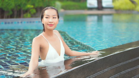 Beautiful-young-lady-kneeling-in-swimming-pool-touching-her-wet-long-hair,-looking-around-and-smilling