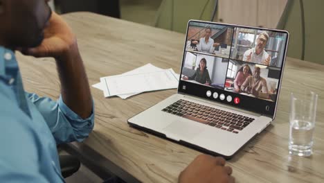 hombre afroamericano usando una computadora portátil para una llamada de video, con diversos colegas de negocios en la pantalla