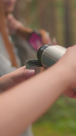 Friends takes break in lush forest, one carefully opens thermos while other gently brushes hair, backpacks rest on fallen tree, capturing relaxed, serene moment as they enjoy nature