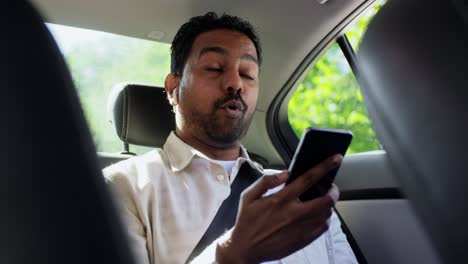 Passenger-with-Earphones-and-Cellphone-in-Taxi-Car.transport,-business-and-technology-concept--happy-smiling-indian-male-passenger-with-wireless-earphones-using-smartphone-on-back-seat-of-taxi-car