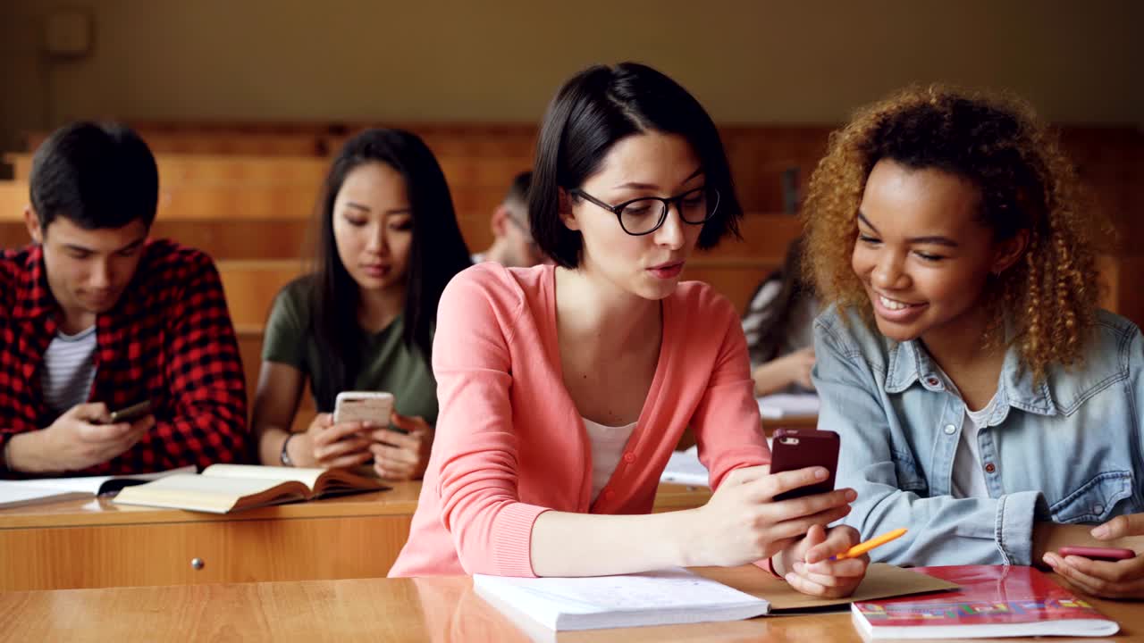 Pretty girls students are using smartphone, watching screen, talking and laughing sitting at desks at university. Social media, internet, millennials and education concept.