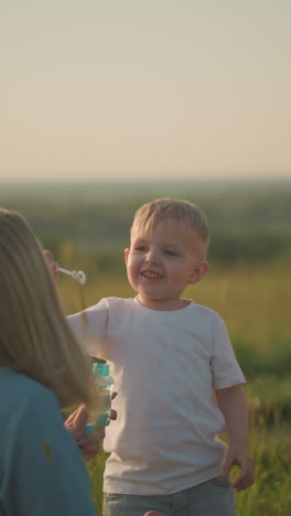 una mujer con un vestido azul se arrodilla en un campo cubierto de hierba al atardecer, sosteniendo una botella de burbujas mientras su hijo menor sopla burbujas de la varita. su hijo mayor, vestido con un traje amarillo, también sopla burbullas