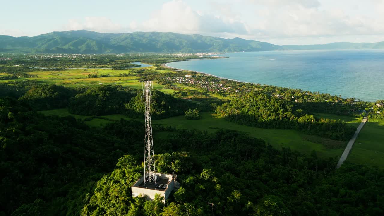 Aerial flyover shot of communication and telecom tower atop lush hill overlooking tropical island villages and coastline - Catanduanes, Philippines