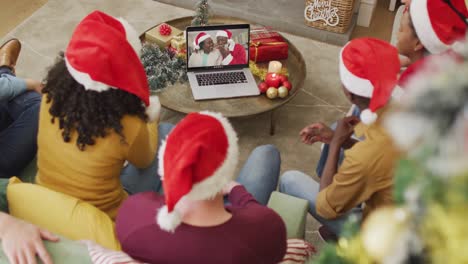 Diverse-family-with-santa-hats-using-laptop-for-christmas-video-call-with-happy-couple-on-screen