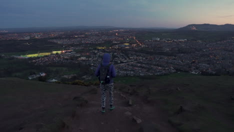 dolly siga la foto de una niña caminando por la montaña arthurs seat en el sendero rocoso por la noche, el anochecer con la ciudad de edimburgo al fondo durante la maravillosa hora azul