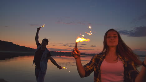 young happy multiethnic couple holding burning sparkling candles and running by the sea during sunset. slow motion shot