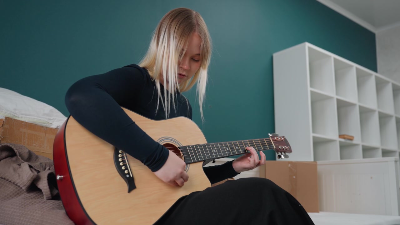 Donna bianca che accorda la chitarra in soggiorno vicino a scaffali non ancora sistemati e scatole di cartone, pratica concentrata, parete color verde acqua brillante, mentre sistema i mobili e allestisce lo studio di casa, energia piena di speranza.