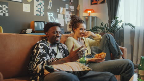 young couple laughing while watching tv and enjoying snacks at home