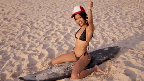 cheerful woman in bikini sitting on surfboard on sand