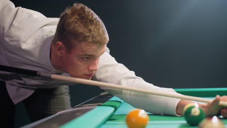 close up of young man with serious face, dressed in white shirt, leaning over green pool table in dimly lit room. gripping cue stick with intense focus, preparing strategic shot with precision