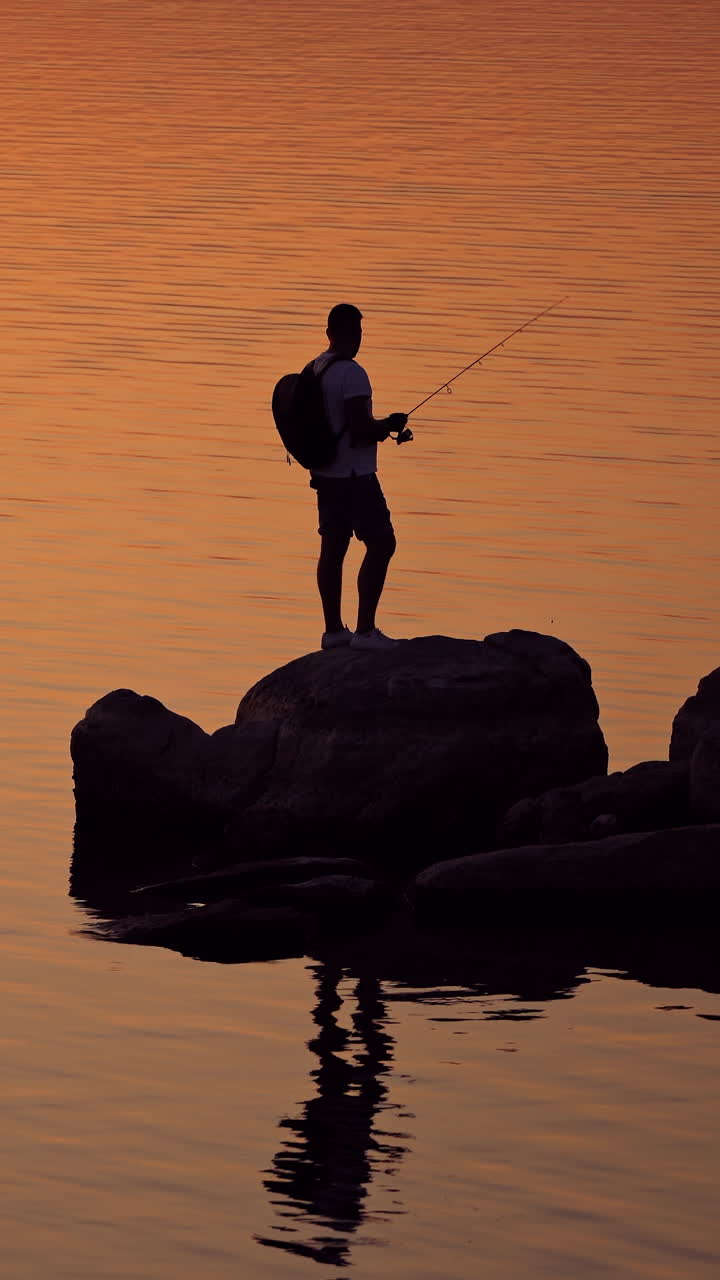 Silhouette of a fisherman fishing on the rock at sunset. Young man with rucksack is standing with fishing rod on a big stone near the river in the evening. Vertical video